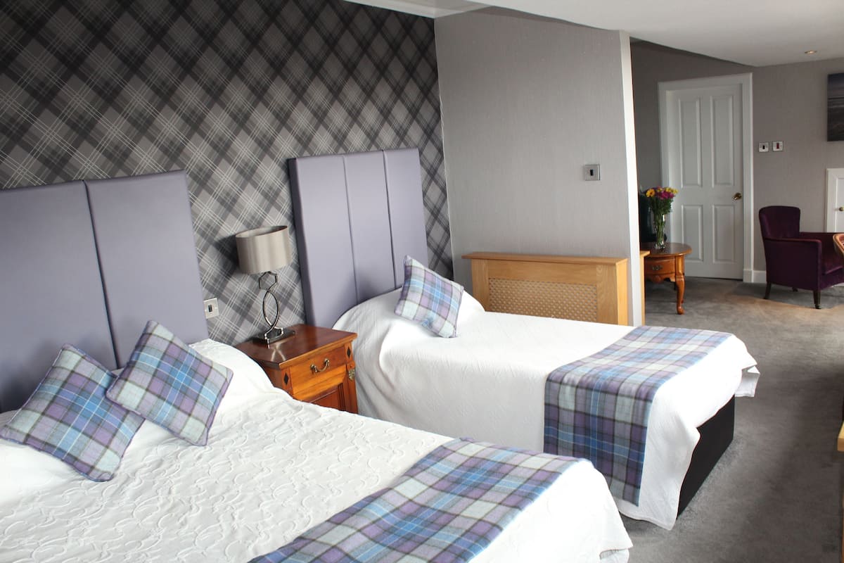 A hotel room with two beds, a wooden nightstand, a lamp, a chair, and a door leading to another room.