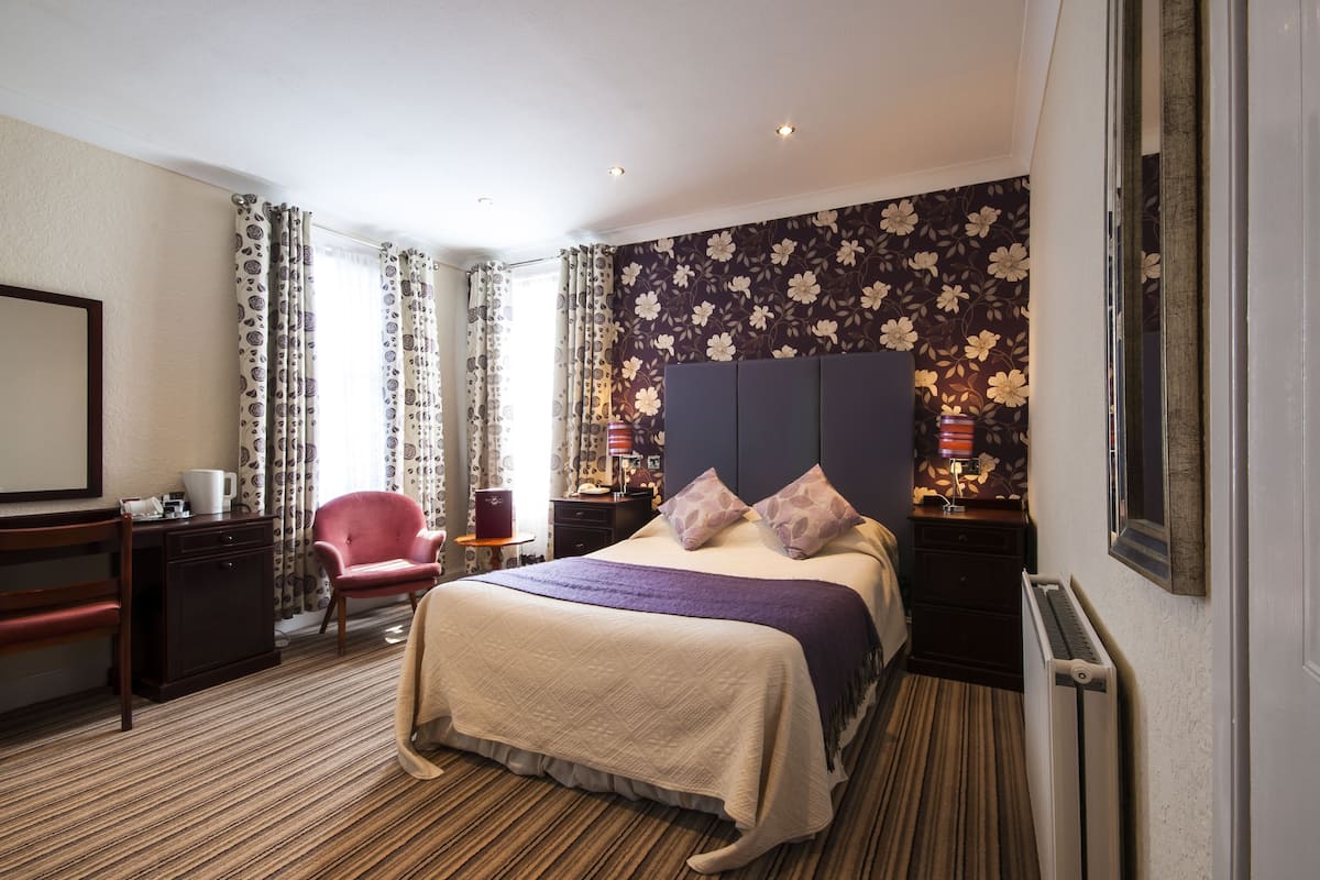 A hotel room with a bed, a desk, a chair, a dresser, and a radiator.