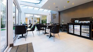 A modern cafe with a glass ceiling, black chairs, and a refrigerator stocked with beverages. - Kensington Prime Hotel (London)