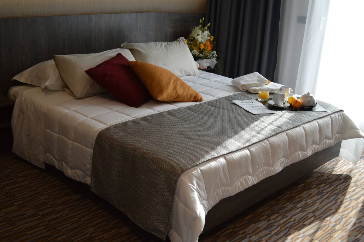 A neatly made bed with a gray and white duvet, a red pillow, and a brown accent pillow. A tray with a teapot, a cup, and a plate of fruit is on the bed.