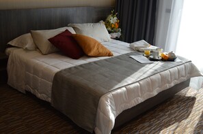 A neatly made bed with a gray and white duvet, a red pillow, and a brown accent pillow. A tray with a teapot, a cup, and a plate of fruit is on the bed.