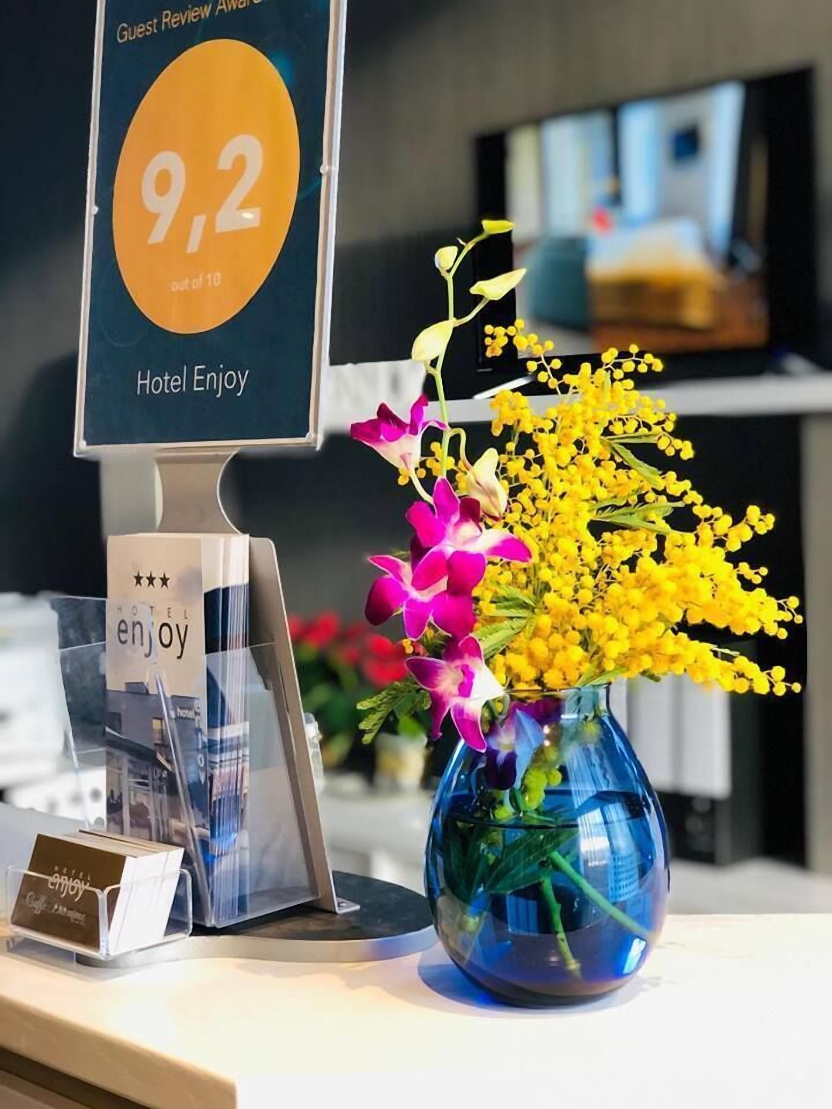 Una reception hotel con un display digitale per le recensioni degli ospiti, un vaso di fiori e un porta cartelli in plastica trasparente.