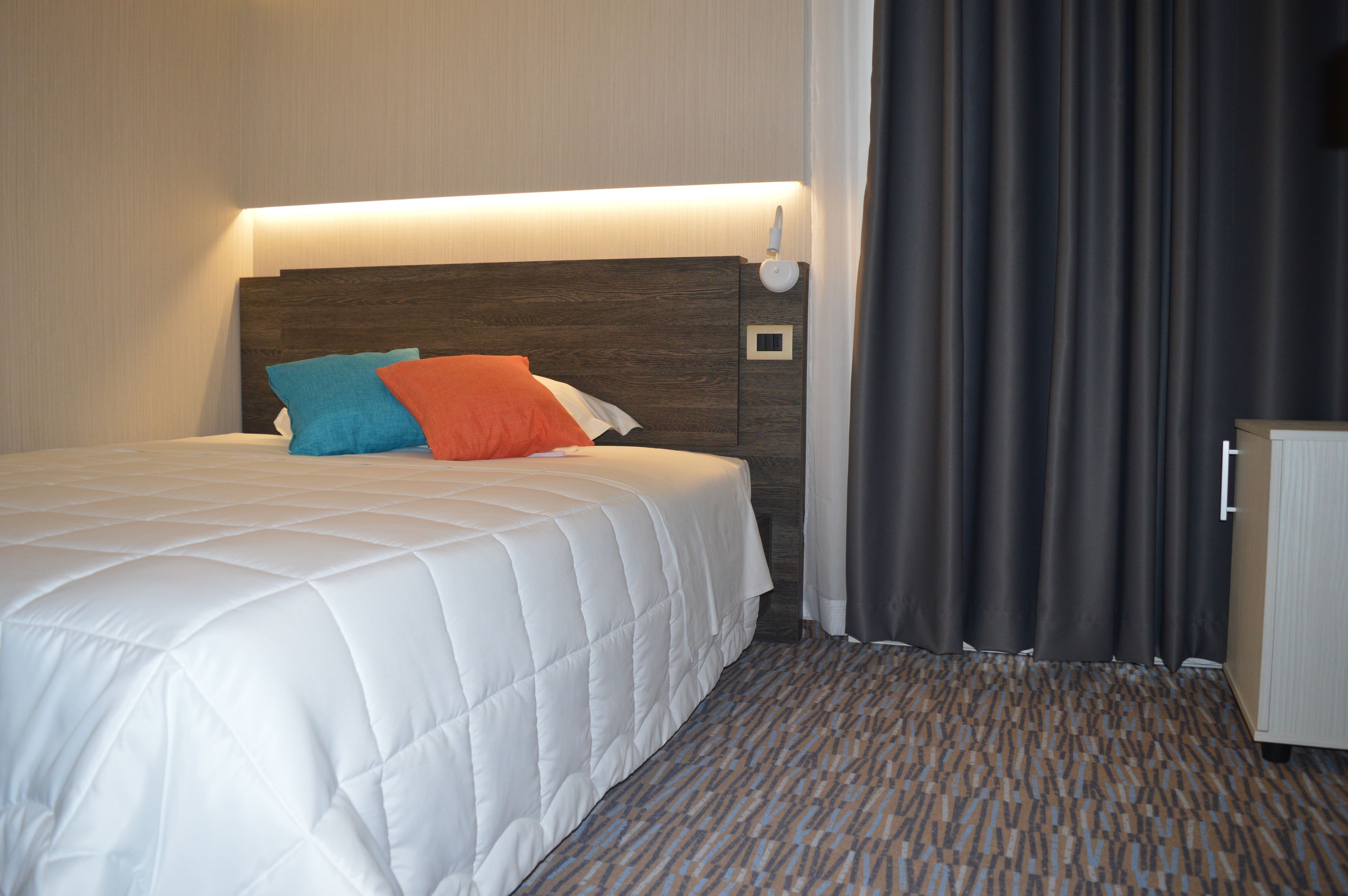 Una habitación de hotel con una cama, cabecero, mesita de noche y cortina.