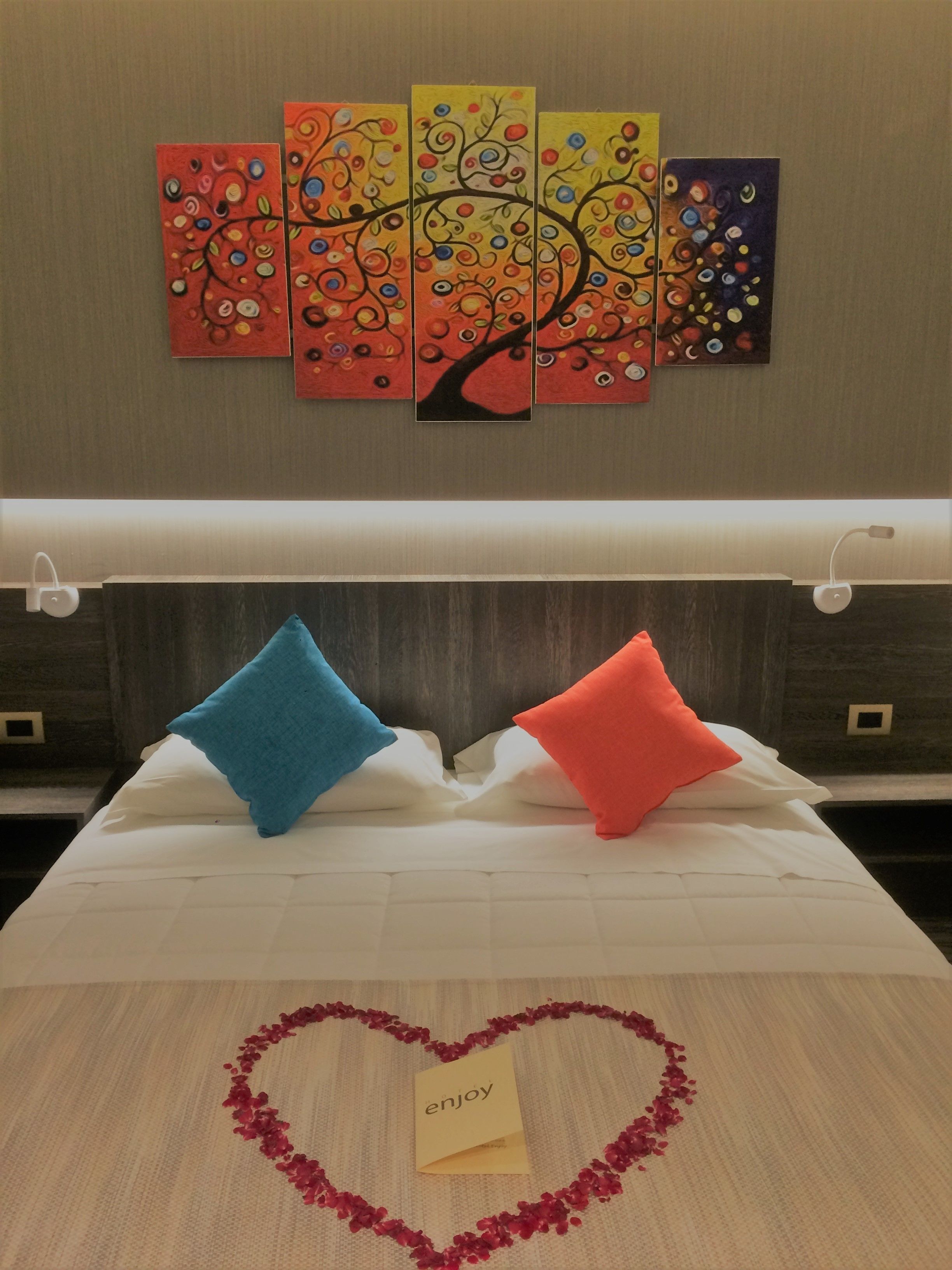 A bed with two pillows, a heart-shaped arrangement of rose petals, and a note saying "enjoy" on a hotel room wall.
