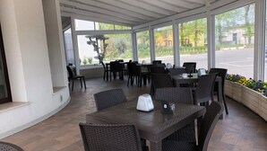 En overdækket udendørs siddeplads med fletstole og borde, store vinduer og udsigt til gaden.