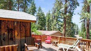 A wooden deck with outdoor furniture, including chairs and a lounge chair, surrounded by tall pine trees.