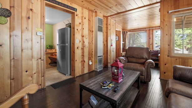 A cabin interior with wooden walls, a dining table with remote controls, a leather sofa, and a refrigerator.