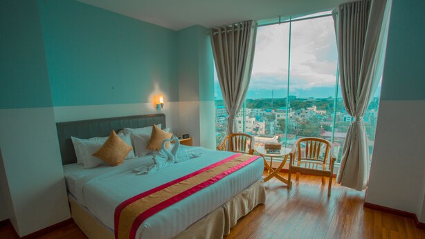 A hotel room with a large bed, a wooden floor, a city view through the window, and a small table with a chair. - Gold Leaf Hotel (Mandalay)