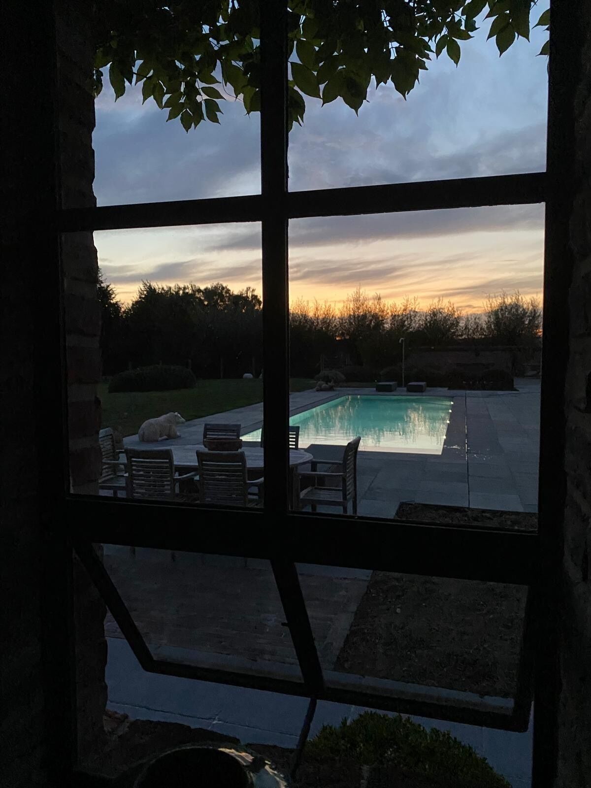 Vista pela janela de uma área de piscina com móveis de exterior e uma parede de pedra.