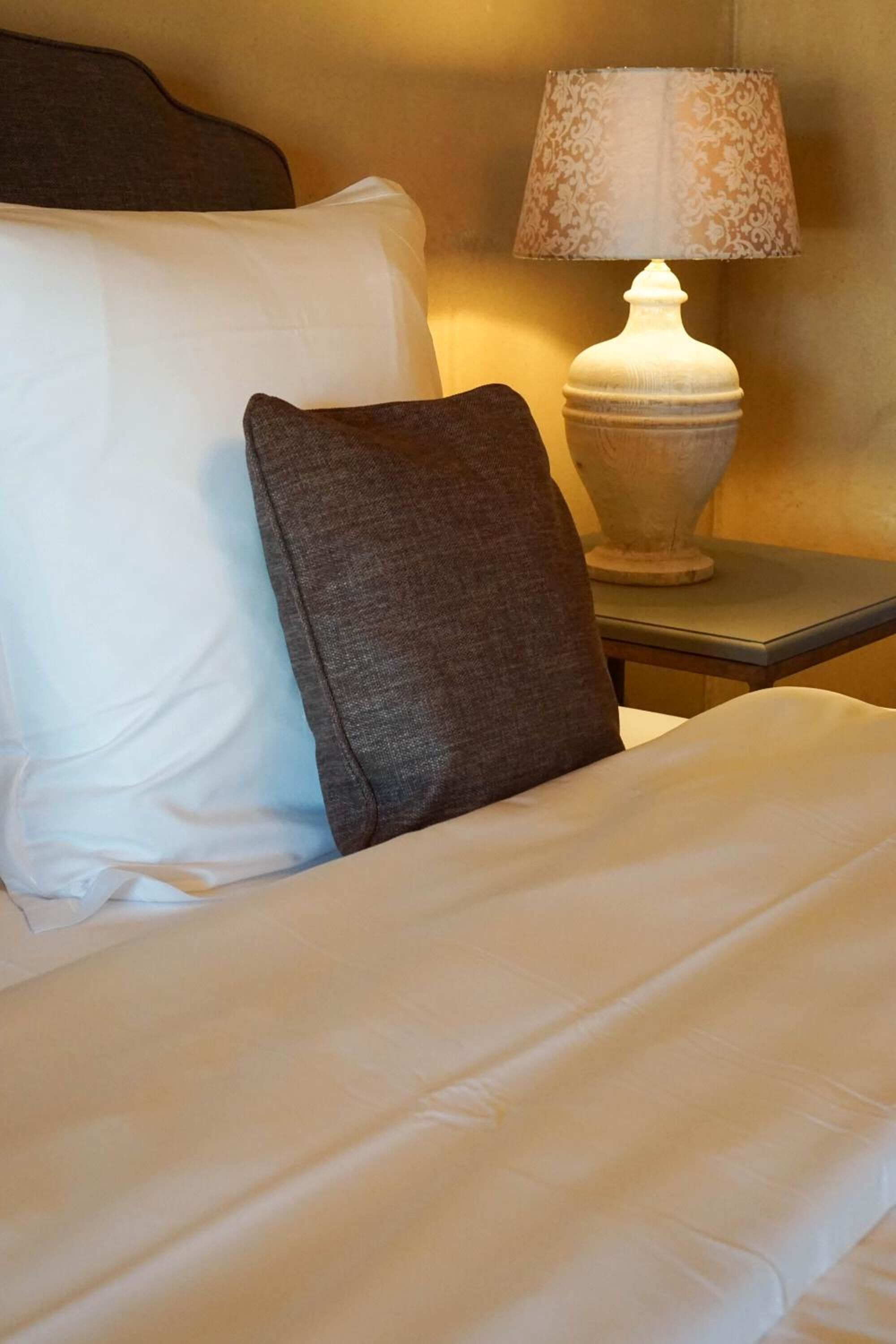 Un letto rifatto con cuscini bianchi e grigio scuro, un piumone bianco e un comodino con una lampada.
