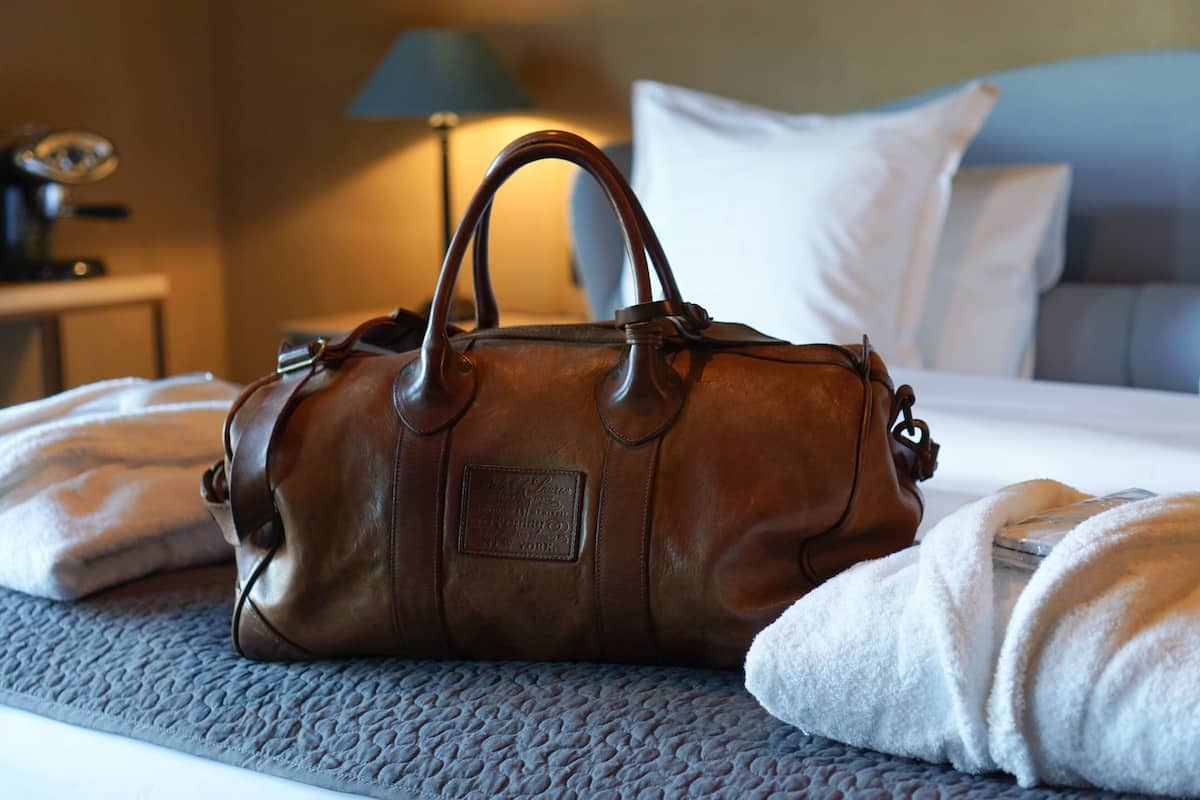 Un bolso de cuero marrón sobre una cama con cubrecama acolchada, una lámpara y una almohada al fondo.