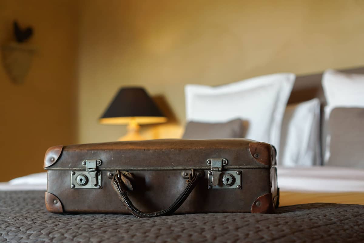 Una maleta marrón sobre una cama, con una lámpara y almohadas al fondo.