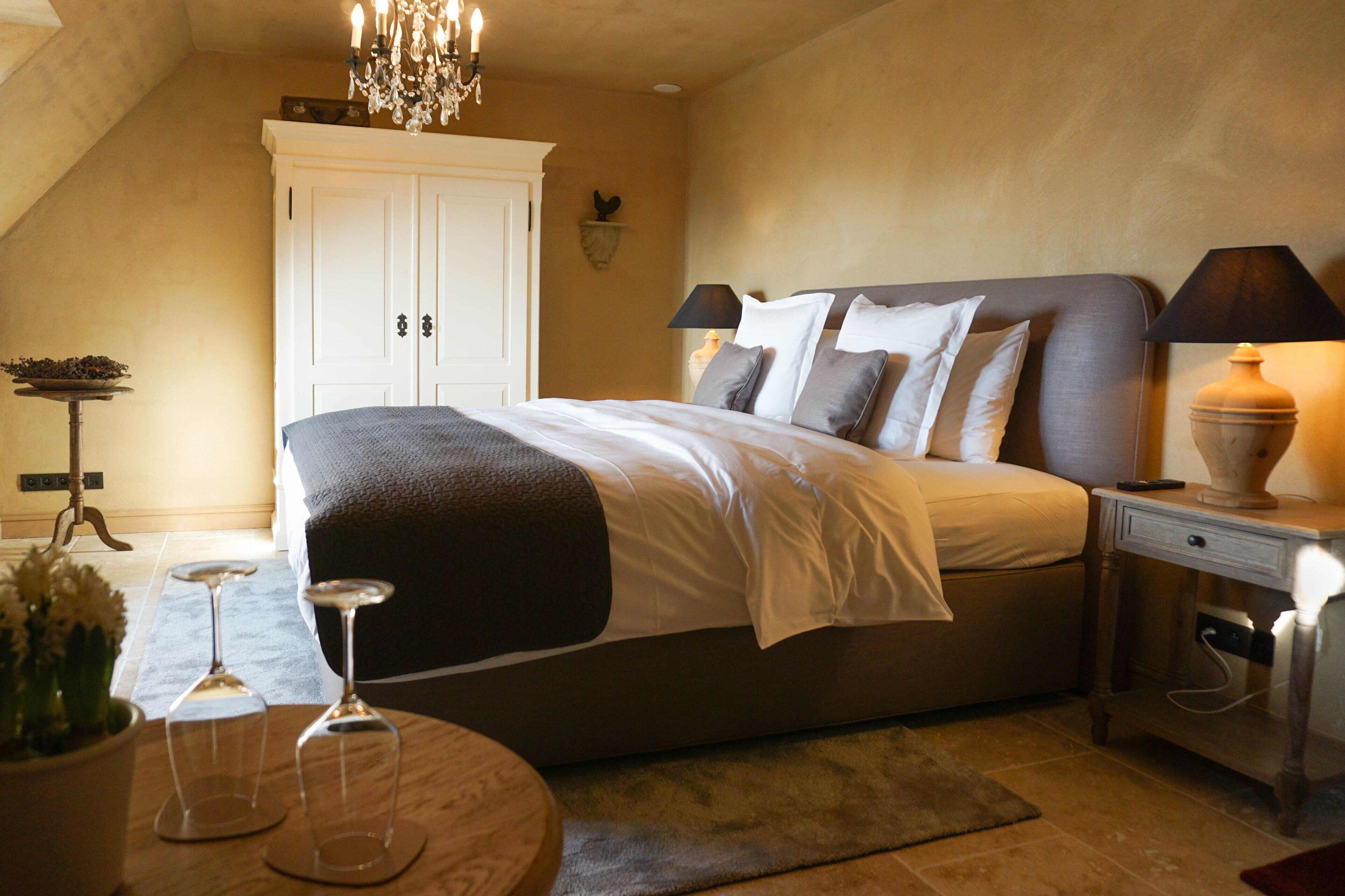 A bedroom with a large bed, bedside lamps, a chandelier, and a small table with wine glasses.