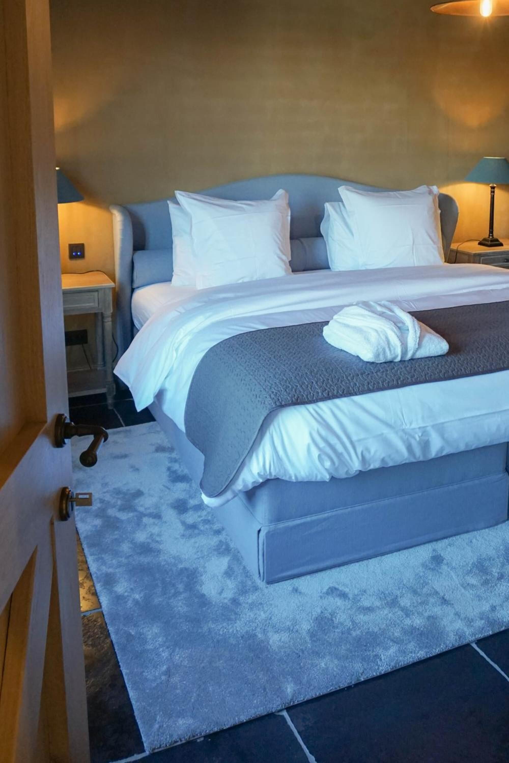 A neatly made bed with white linens and a grey blanket, a bedside table with a lamp, and a wooden door with a handle.
