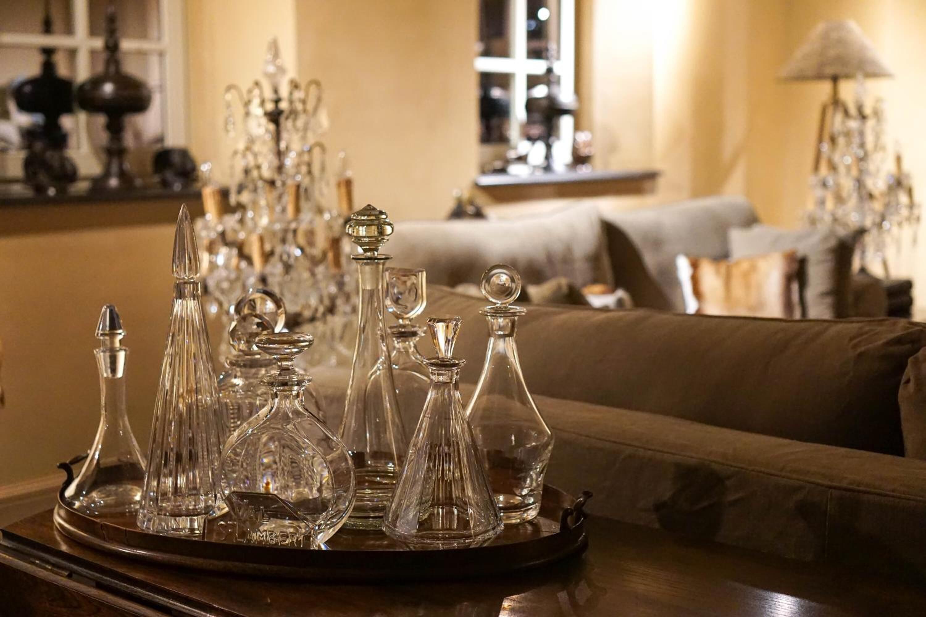 A tray of clear glass decanters on a wooden table in a warmly lit room with a sofa and a lamp in the background.