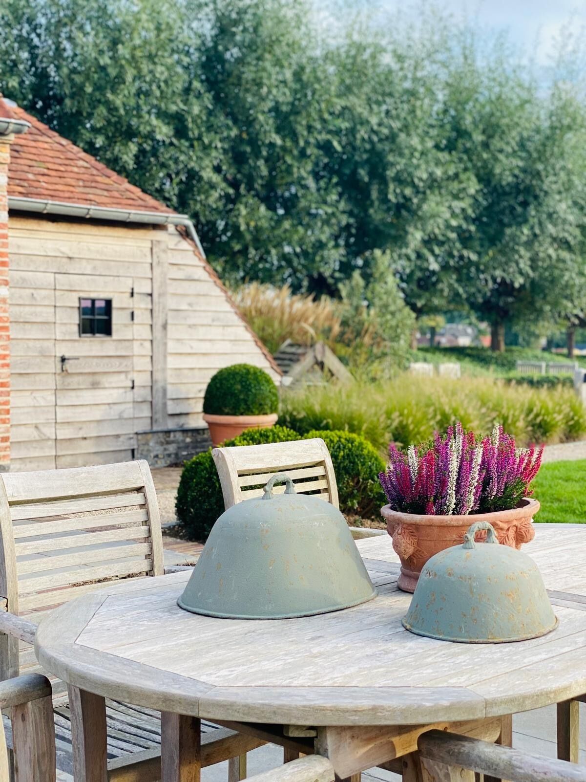 A round wooden table with two metal bell-shaped covers, a wooden chair, and a potted plant with purple flowers.