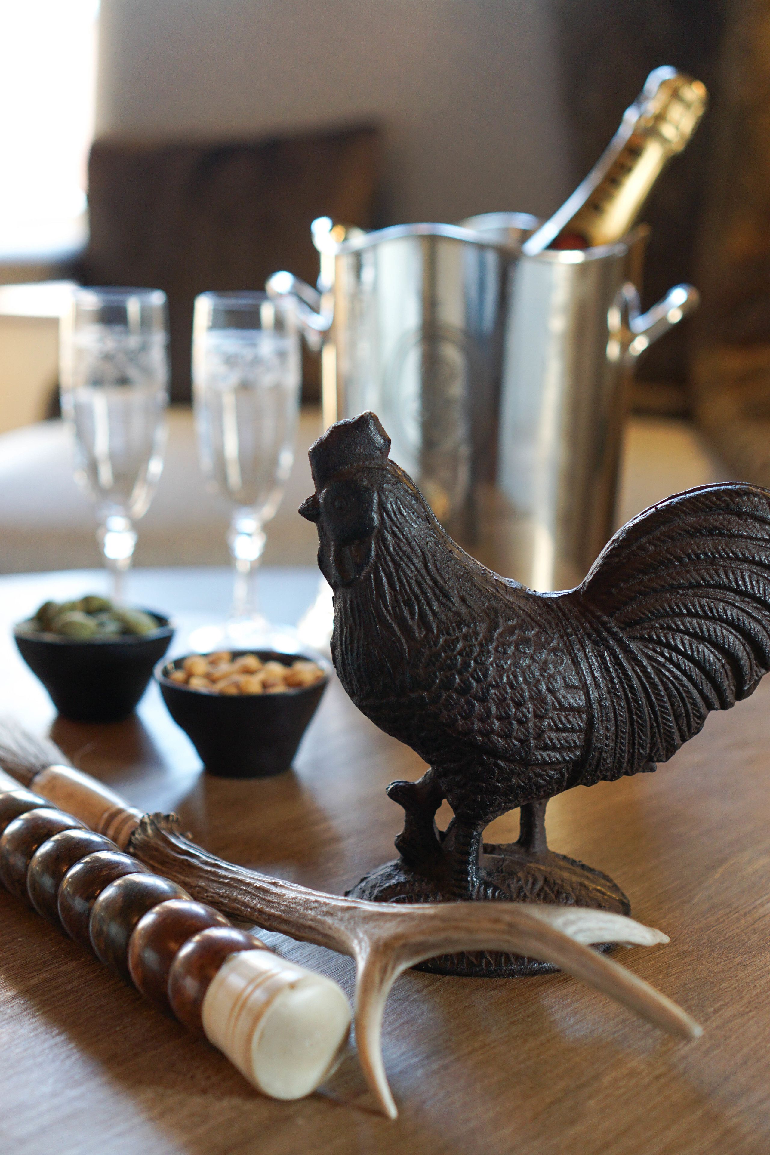 A wooden table with a decorative rooster, a bottle of champagne, and two small bowls of snacks.