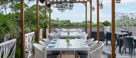 Un espacio de comedor al aire libre con mesas y sillas, ubicado bajo una pérgola de madera con faroles colgantes.