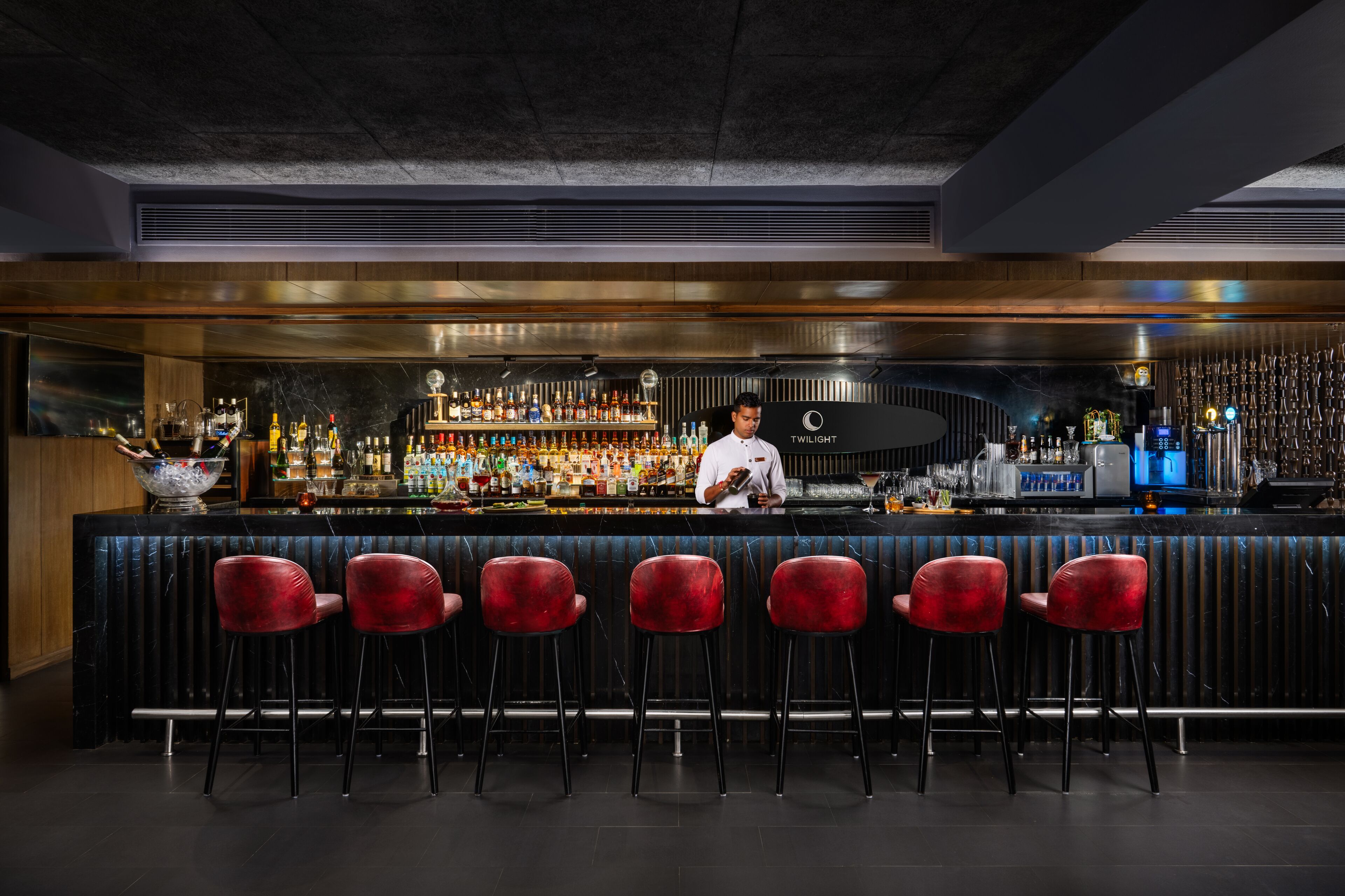 Un bar avec un barman, une variété de bouteilles et des tabourets rouges.