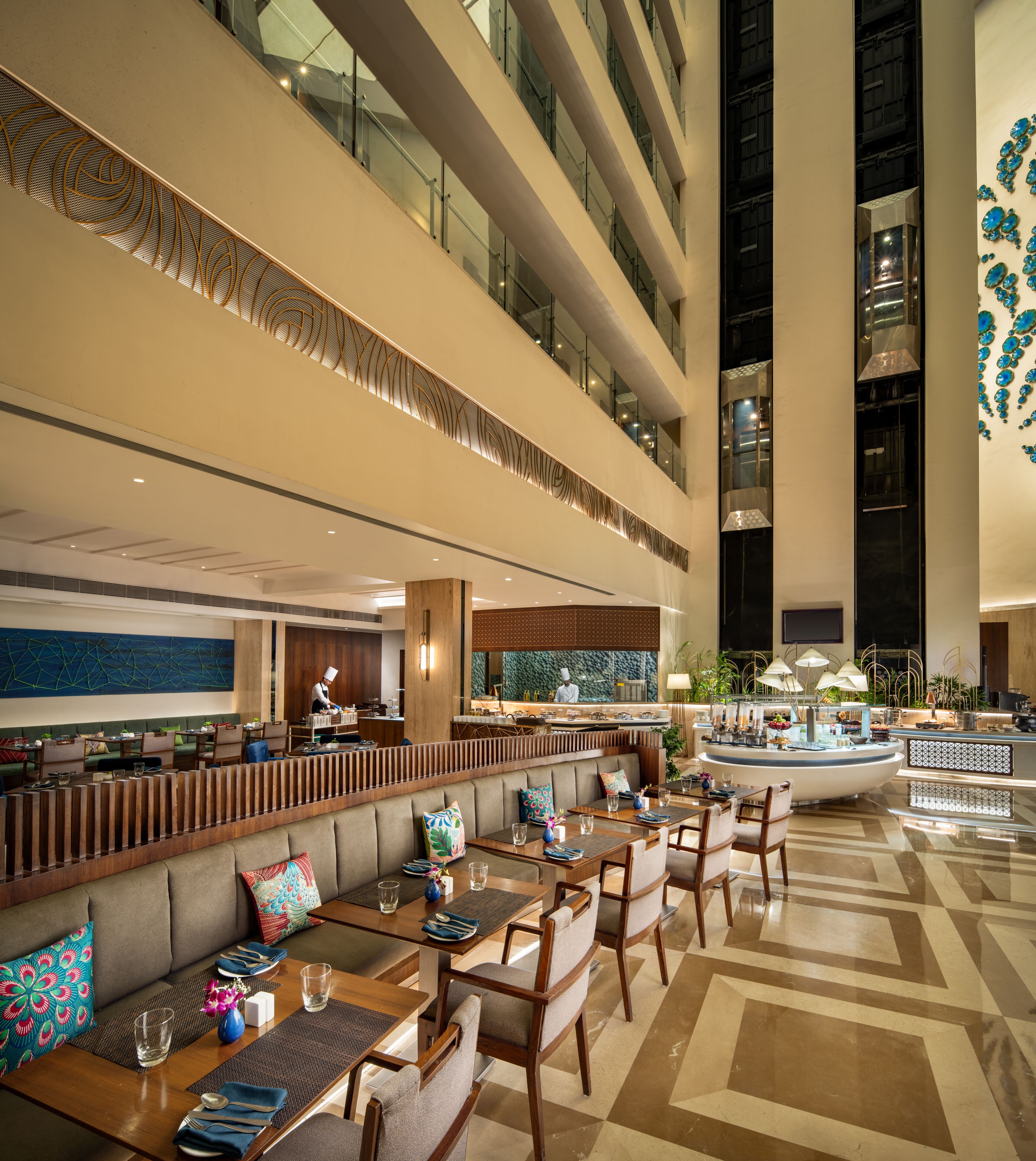 A modern hotel lobby with multiple dining areas, a central bar, and a high ceiling with decorative lighting.