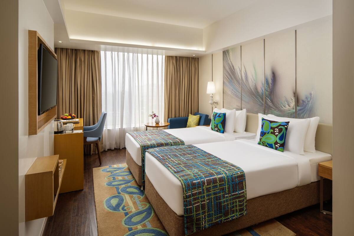 Habitación de hotel con dos camas, un televisor, un escritorio con lámpara y un ventanal con cortinas.