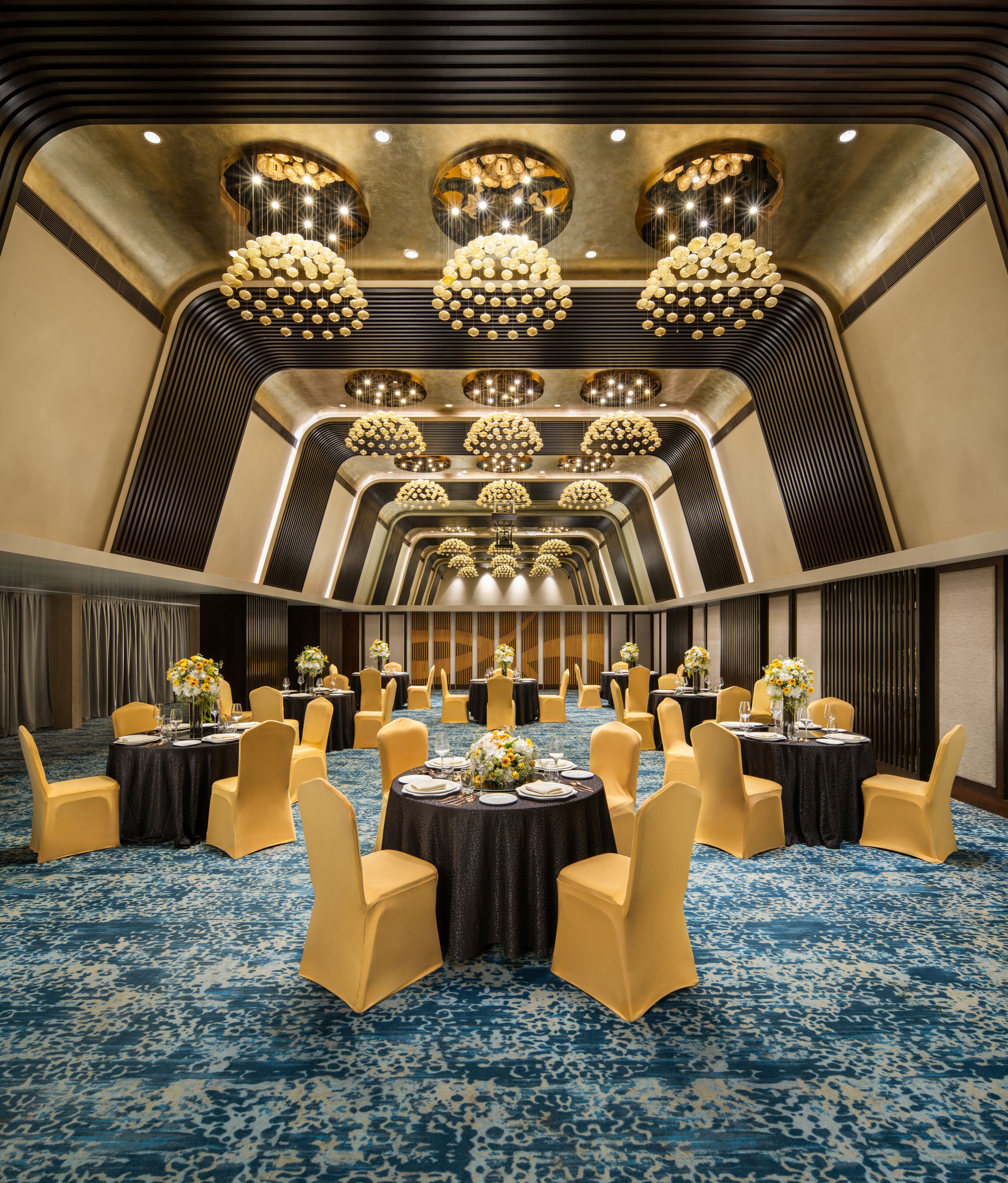 A banquet hall with round tables, yellow chairs, and a blue patterned carpet.
