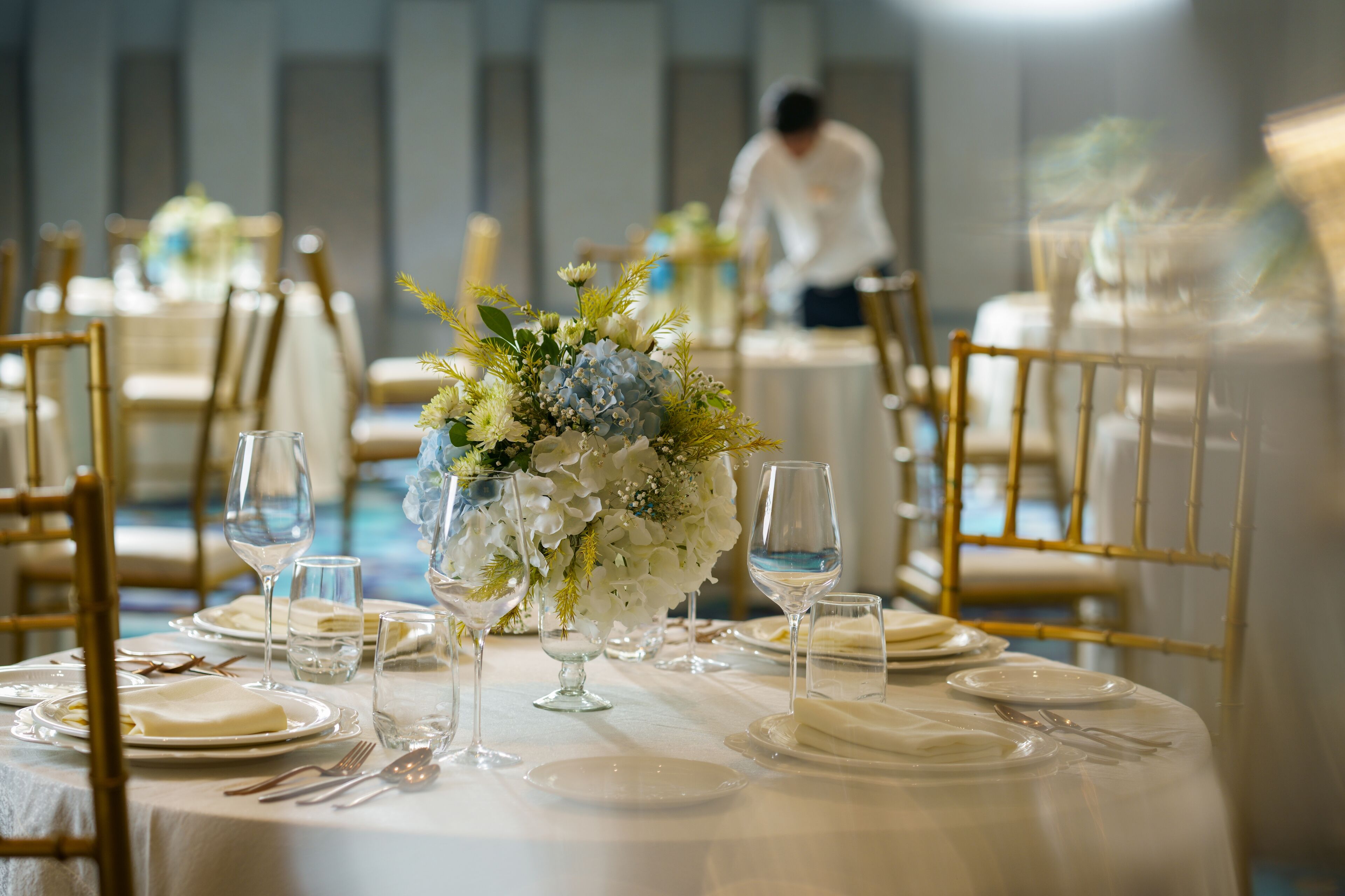 A dining table set with white tablecloths, plates, and wine glasses, surrounded by gold chairs.