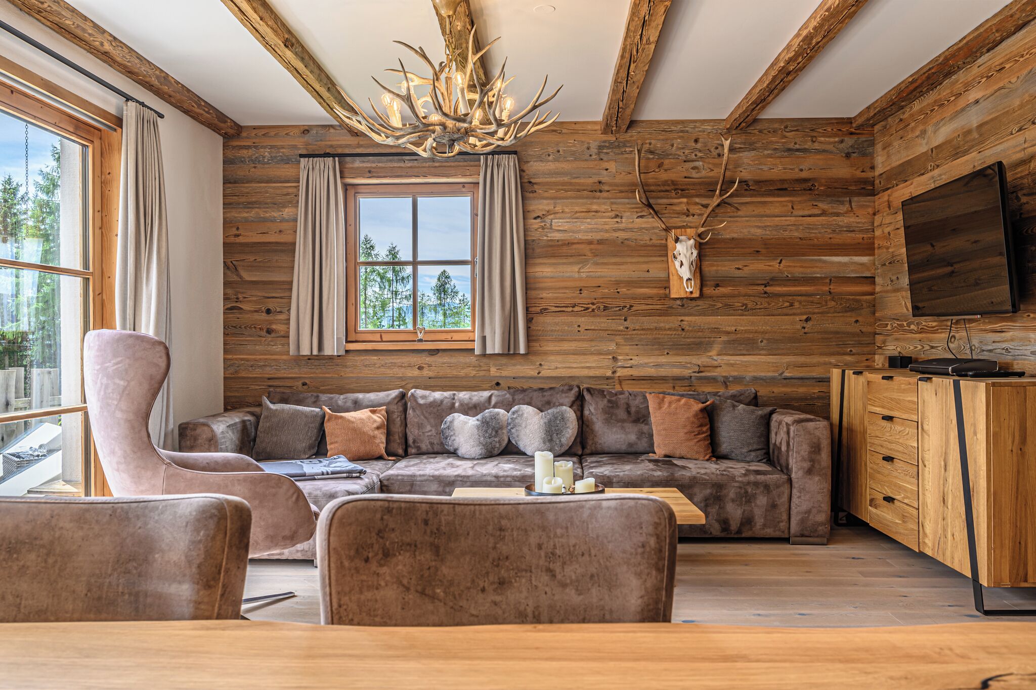 A rustic cabin interior with a large sofa, a plush armchair, a wooden cabinet, and a mounted deer head.