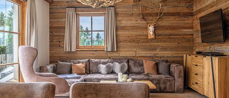 A rustic cabin interior with a large sofa, a plush armchair, a wooden cabinet, and a mounted deer head.