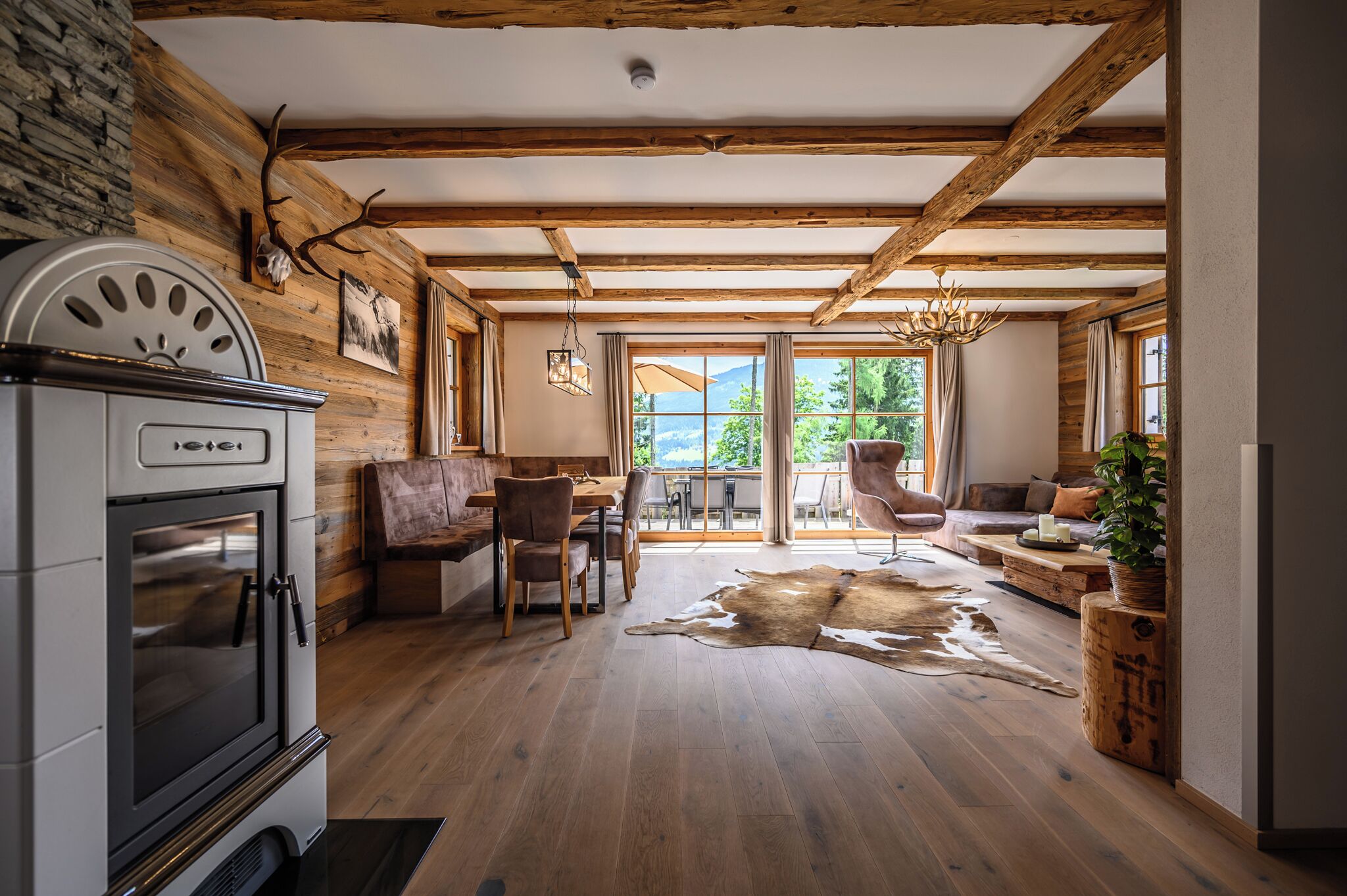 A rustic interior with exposed wooden beams, a stone wall, a fireplace, a dining area, and a large window offering a view of the outdoors.