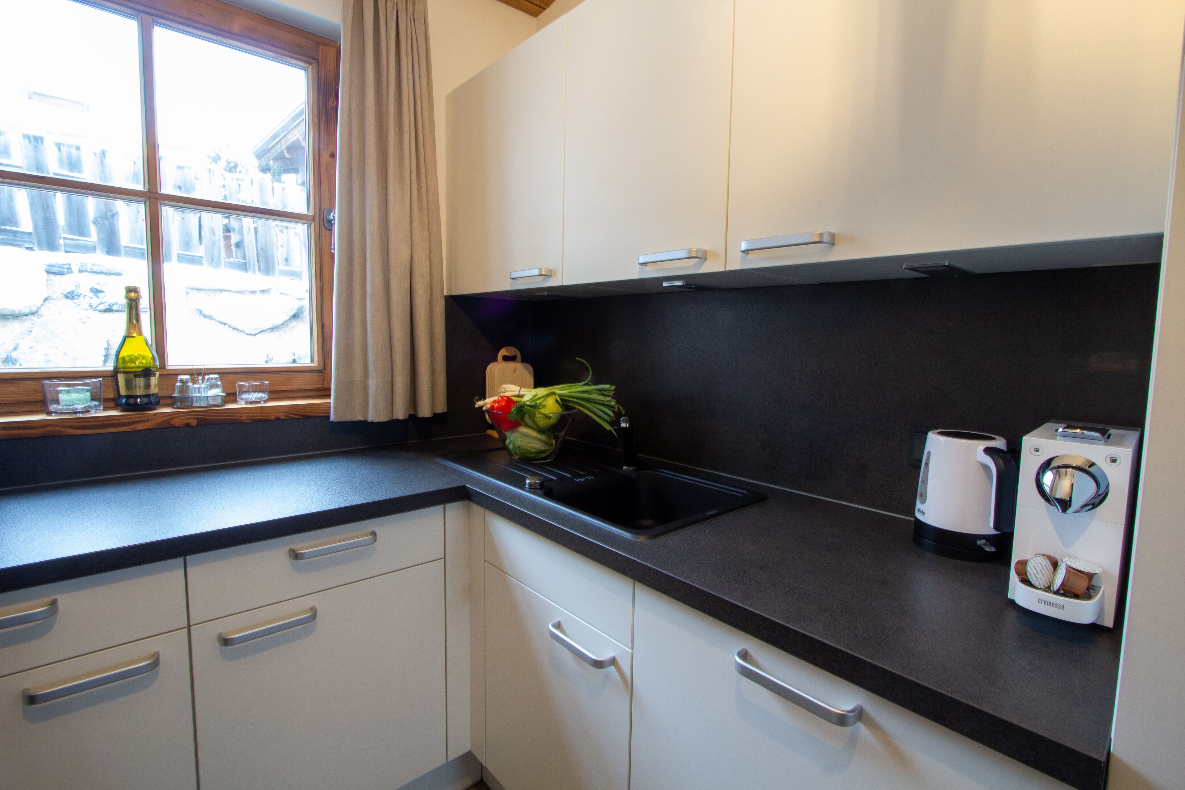 Una cocina moderna con armarios blancos, encimera negra, una ventana con cortinas y una cafetera.