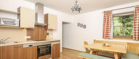 Una cocina compacta con armarios de madera, un microondas integrado y una estufa. Hay una zona de comedor con una mesa y un banco de madera, y una ventana con cortinas.