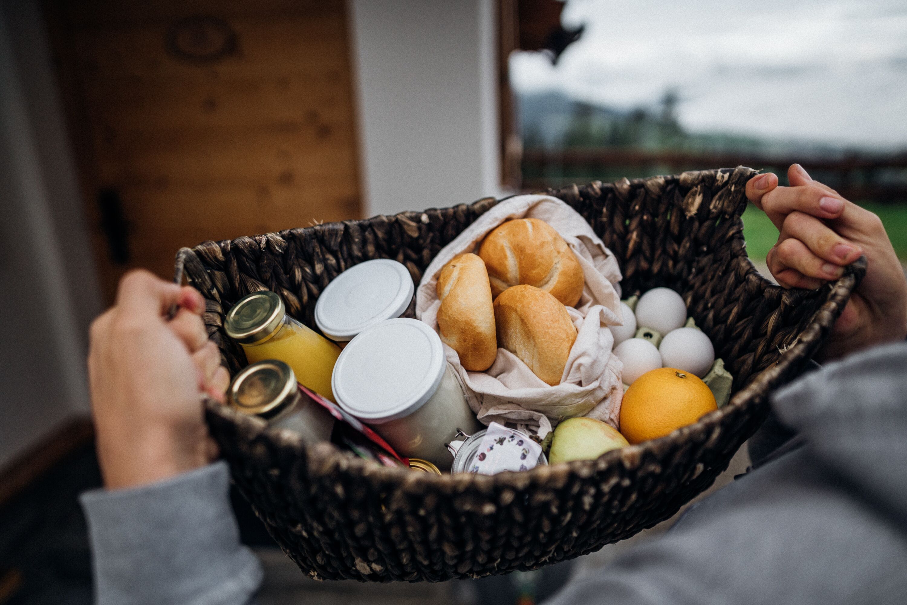 A hand holding a basket containing bread, eggs, and a bottle.