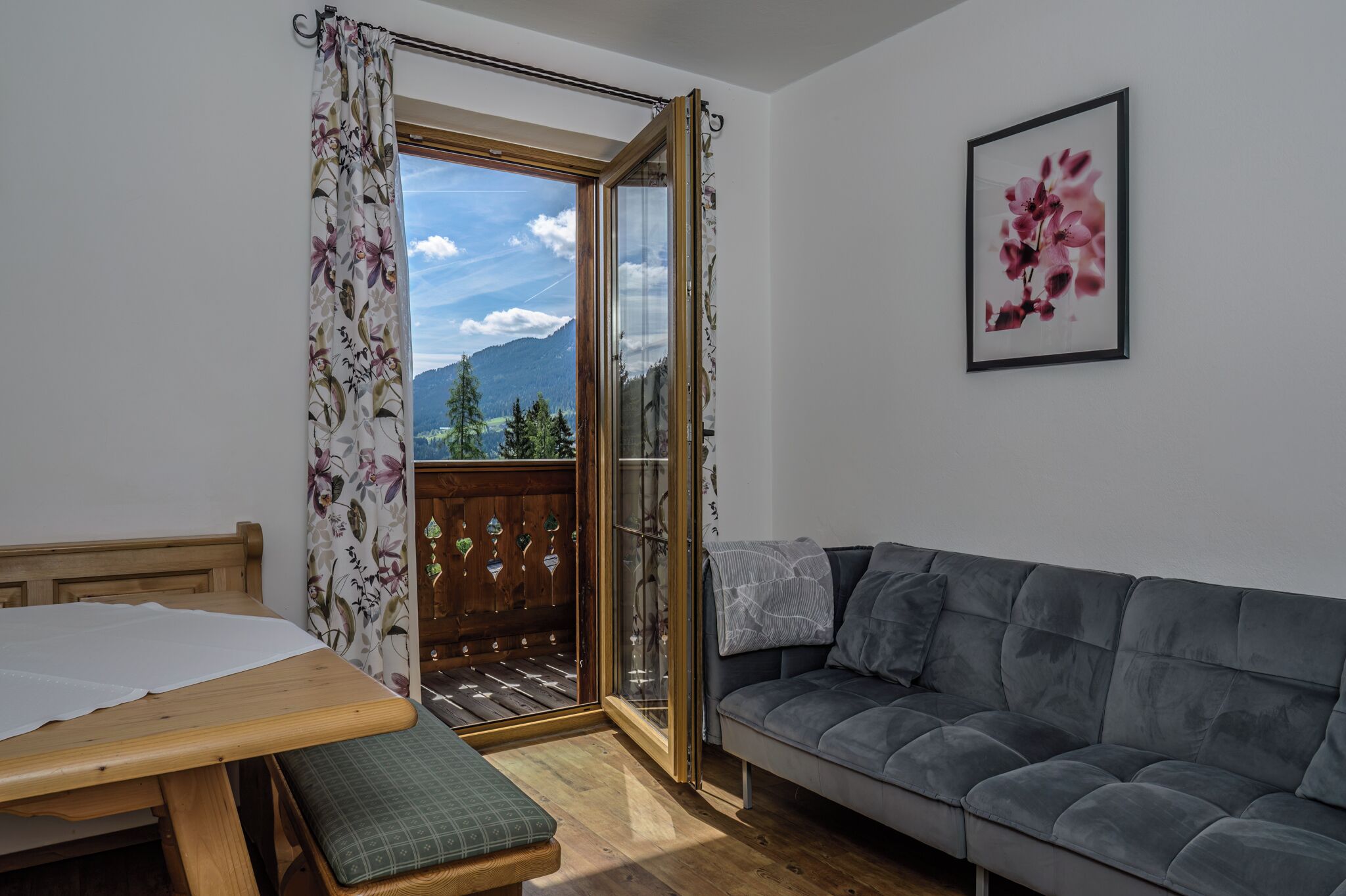 A room with a wooden bed, a grey sofa, a wooden table, and a view of mountains through a glass door.