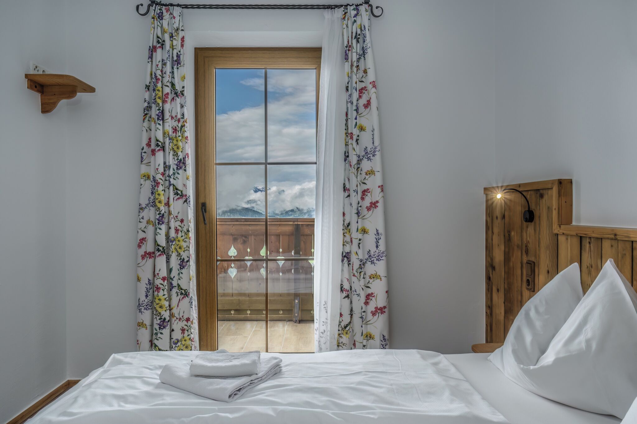 A bedroom with a bed, white bedding, a wooden headboard, a wooden shelf, and a view of mountains through a glass door.