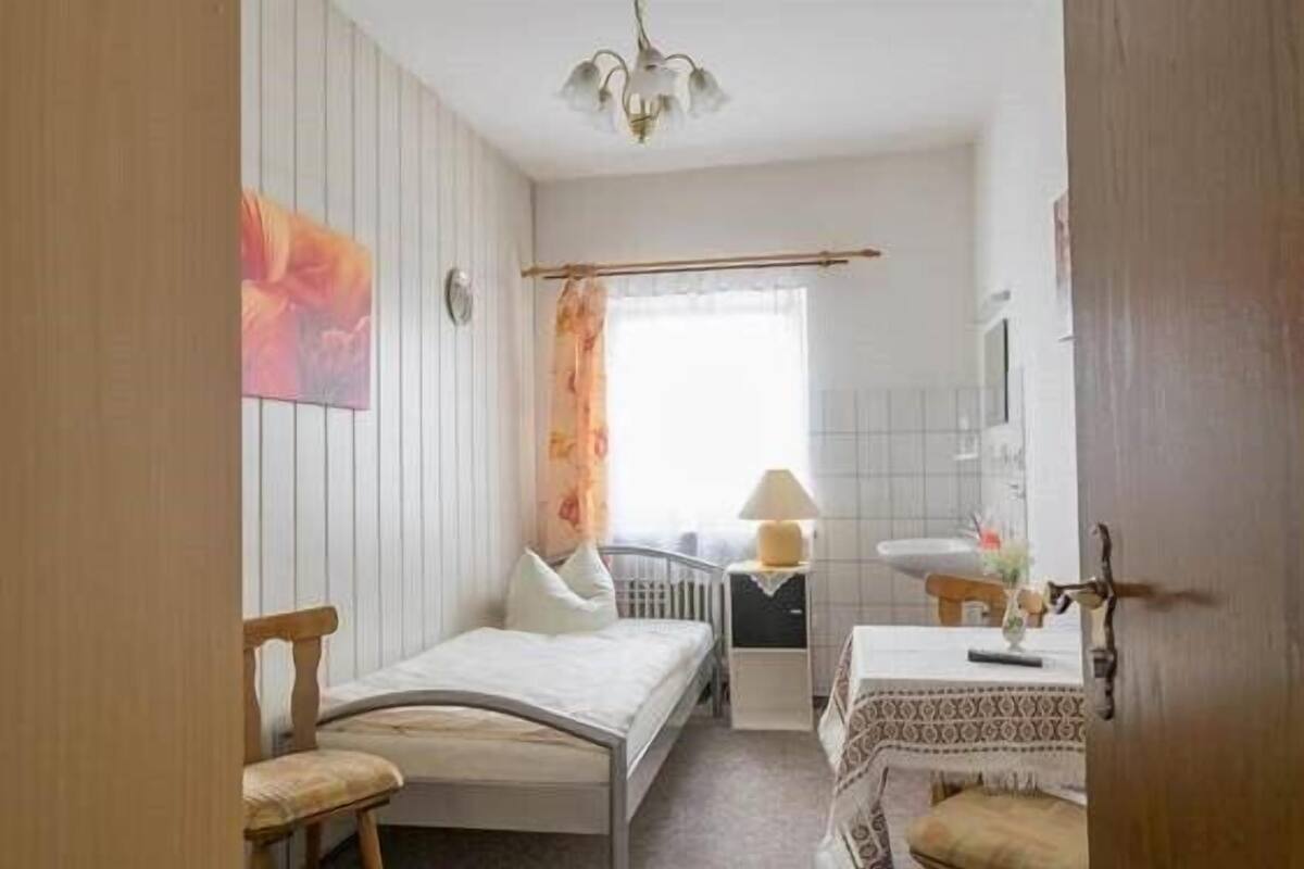 Un dormitorio pequeño y bien iluminado con una cama individual, una mesita de noche, una silla y una ventana con cortinas.