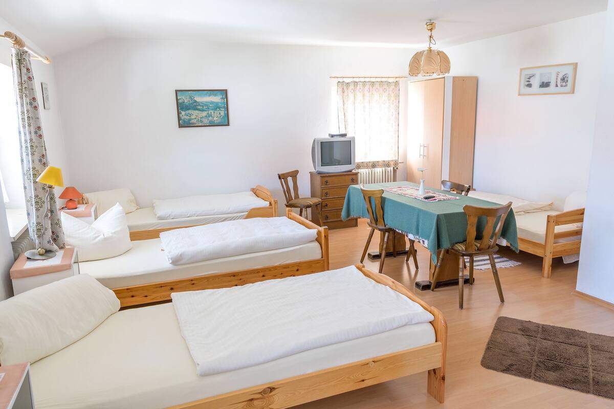 Una habitación con tres camas de madera, televisión, una mesa con sillas y un ventanal con cortinas.
