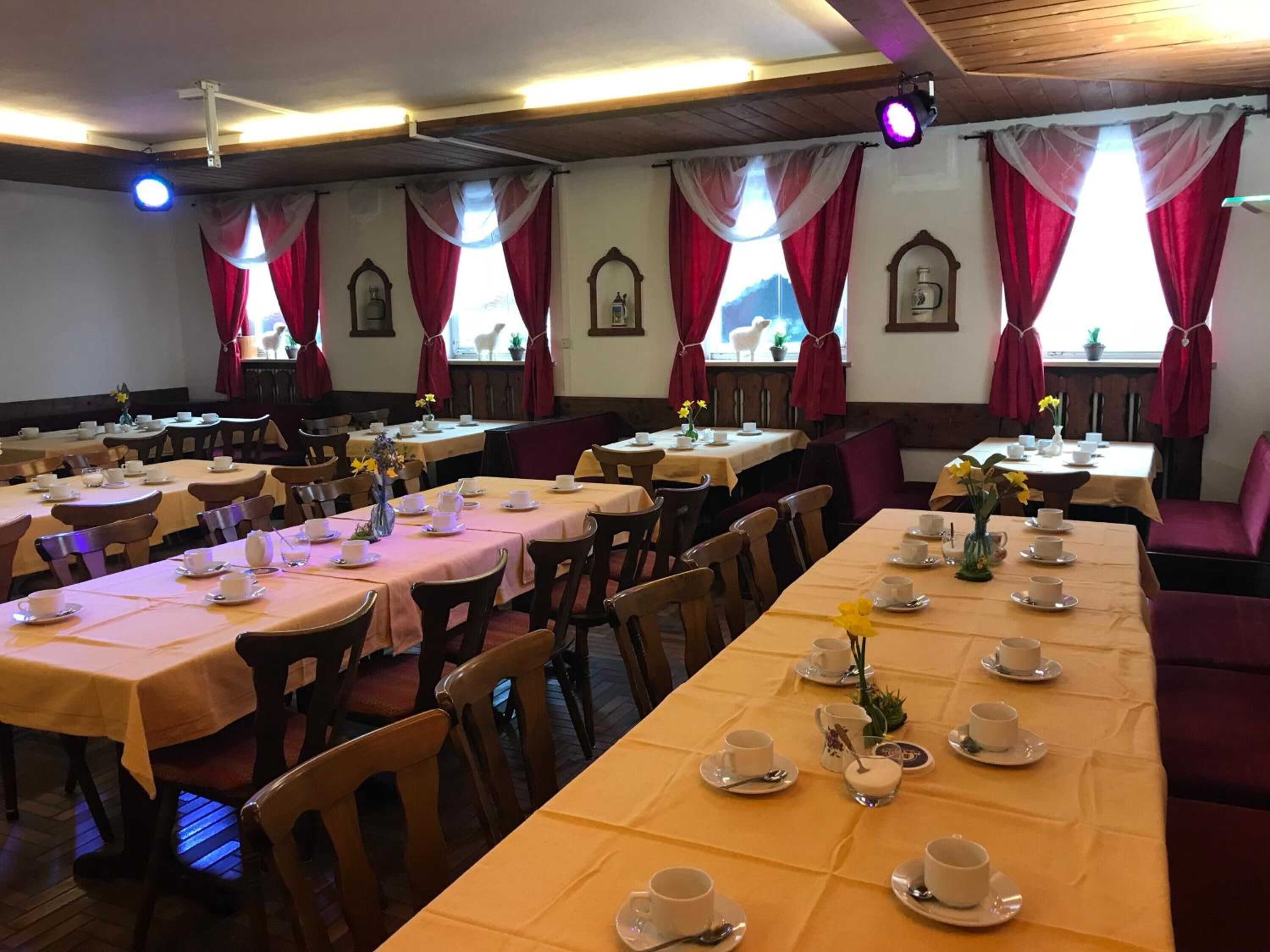 Un comedor con varias mesas dispuestas para una merienda, con tazas y platillos blancos y cortinas rojas.