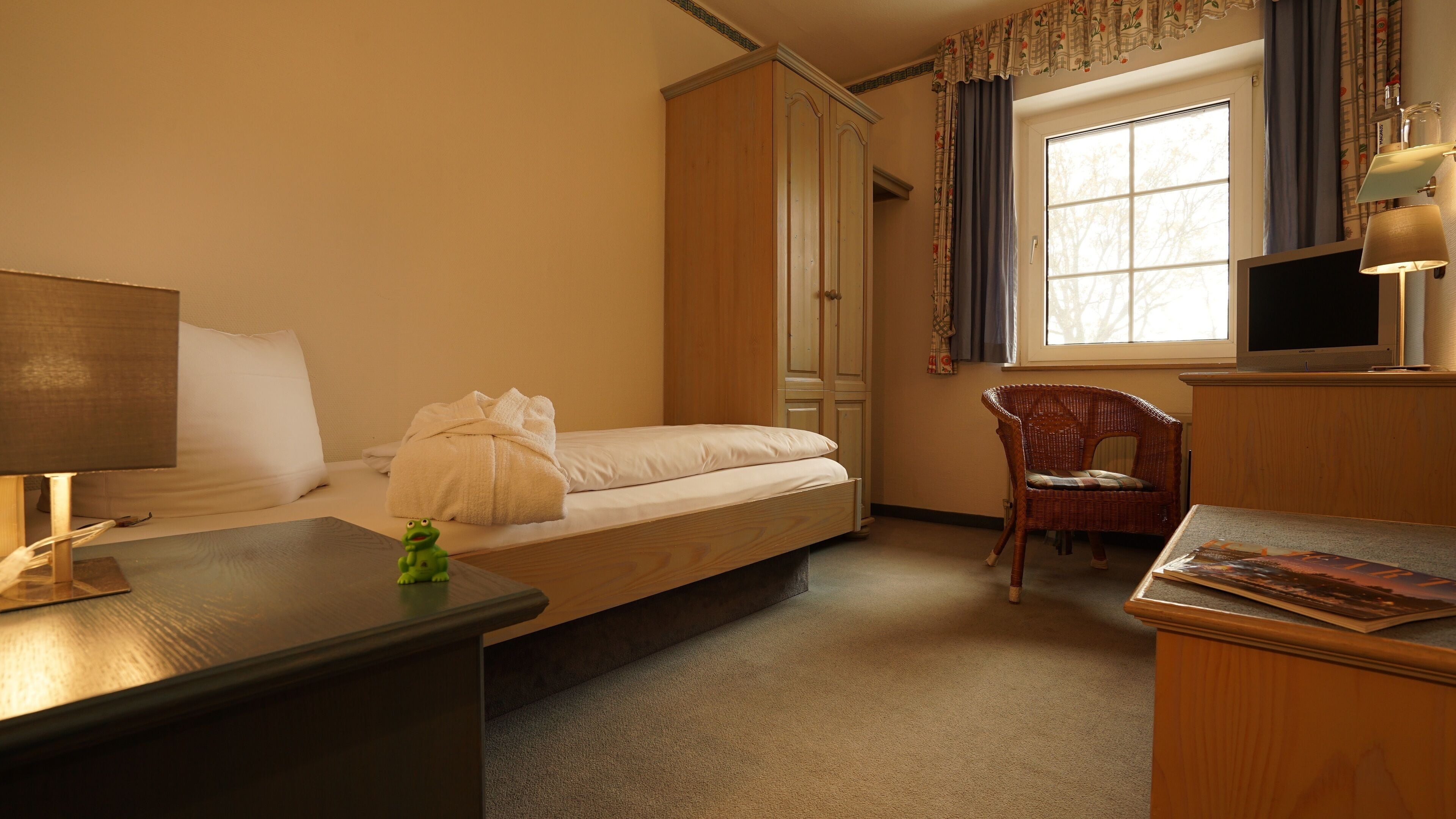 Un dormitorio con una cama individual, un escritorio con lámpara, una silla, un televisor y una ventana con cortinas.