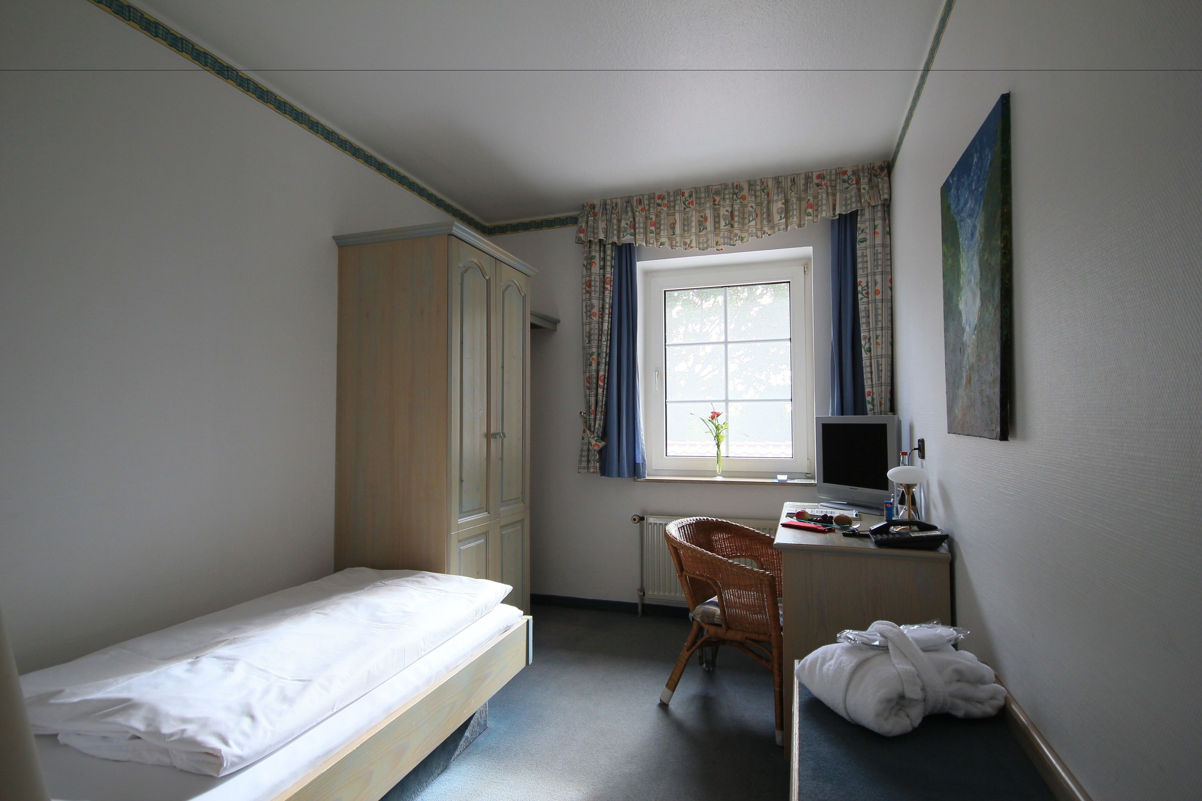 Una habitación pequeña y limpia con una cama individual, un escritorio con silla, un radiador y una ventana con cortinas.