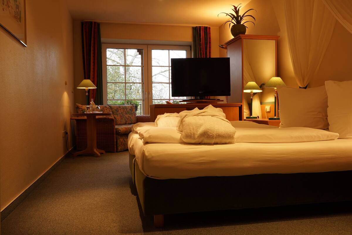 Quarto de hotel com uma cama grande, televisão, escrivaninha com luminária e janela com cortinas.