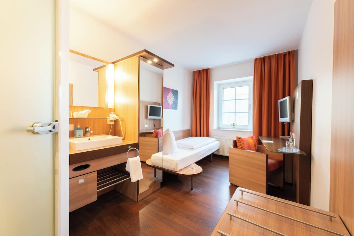 Quarto de hotel moderno com piso de madeira, um espelho grande, cama, escrivaninha, televisão e janela com cortinas.