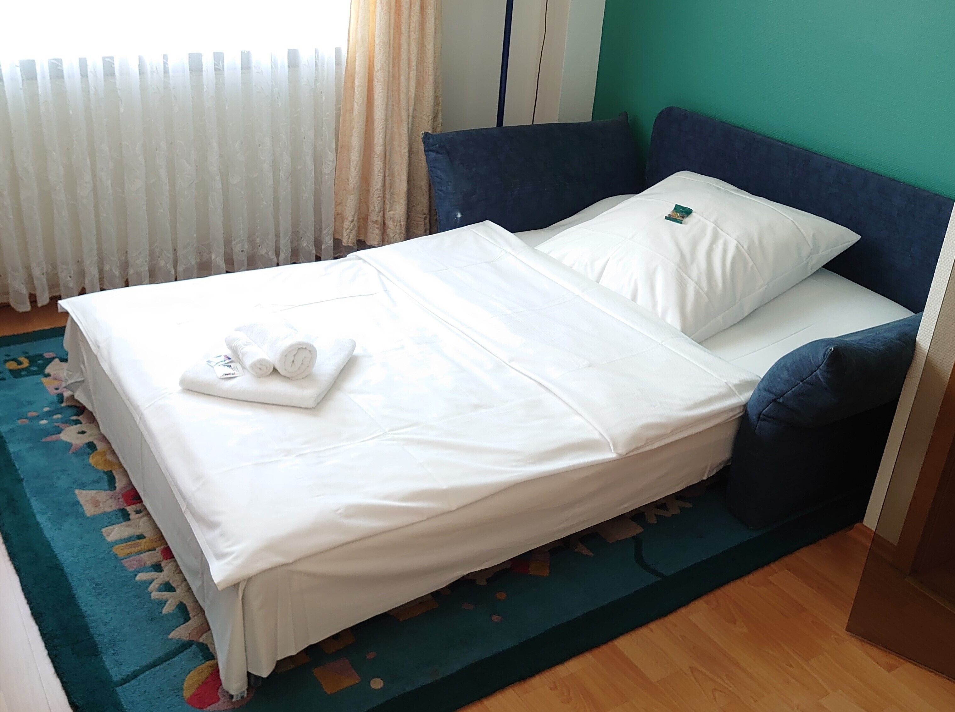 Ein ordentlich bezogenes Bett mit weißen Leinen und aufgerollten Handtüchern, eine wandfarbene Wand in Türkis und ein gemustertes Teppich.