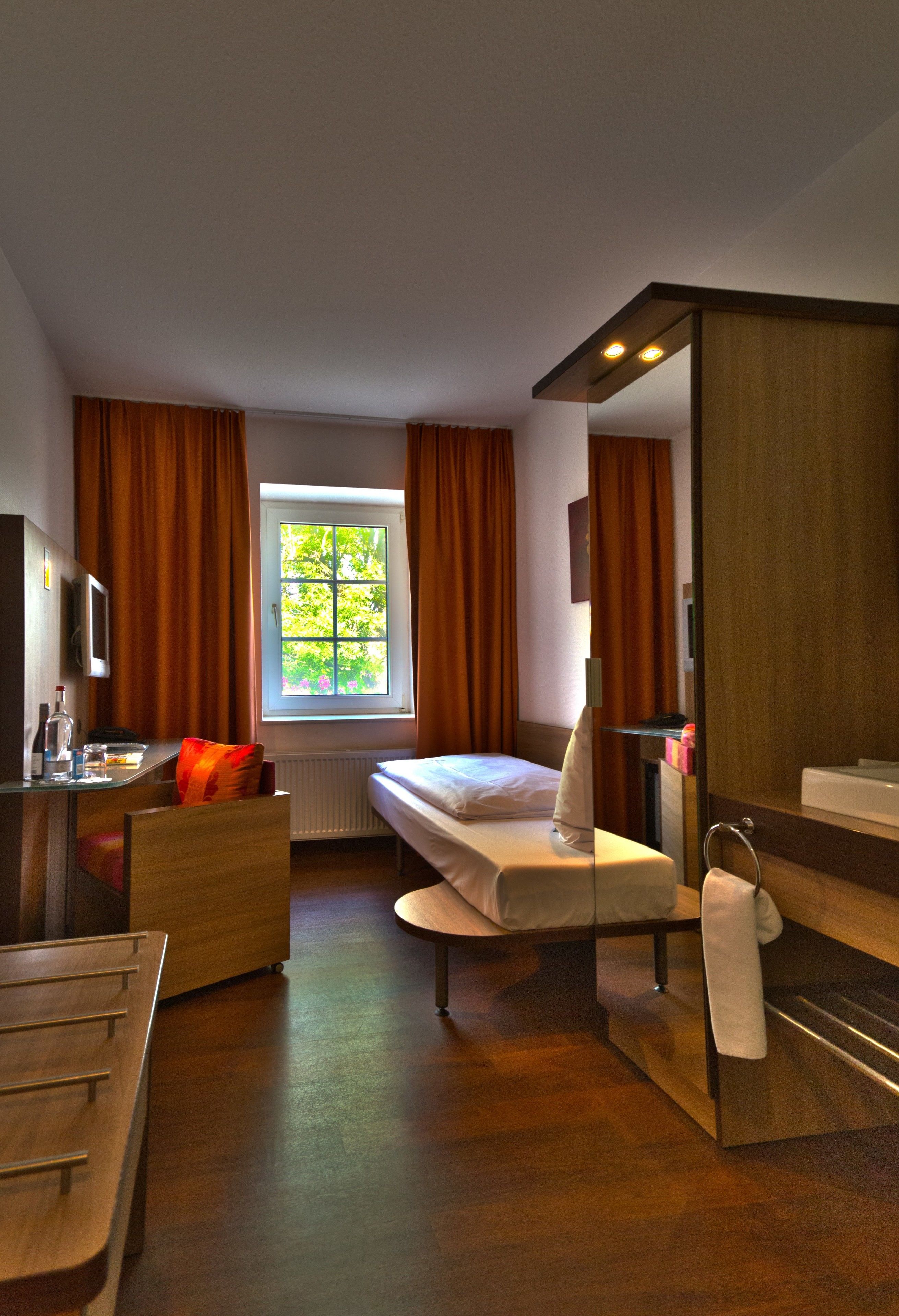 A hotel room with a bed, a desk with a chair, a bathroom with a mirror and sink, and a window with curtains.