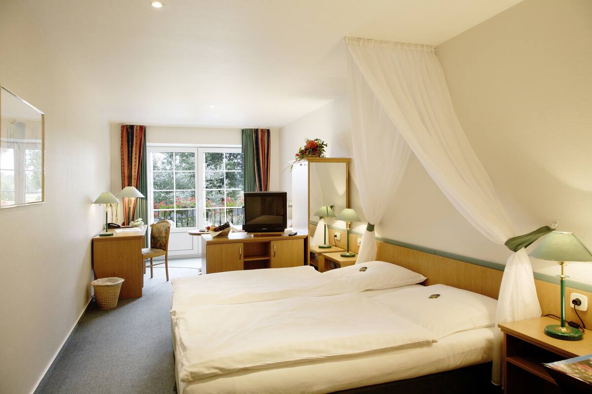 Quarto de hotel com cama, escrivaninha, cadeira, televisão e janela com cortinas.
