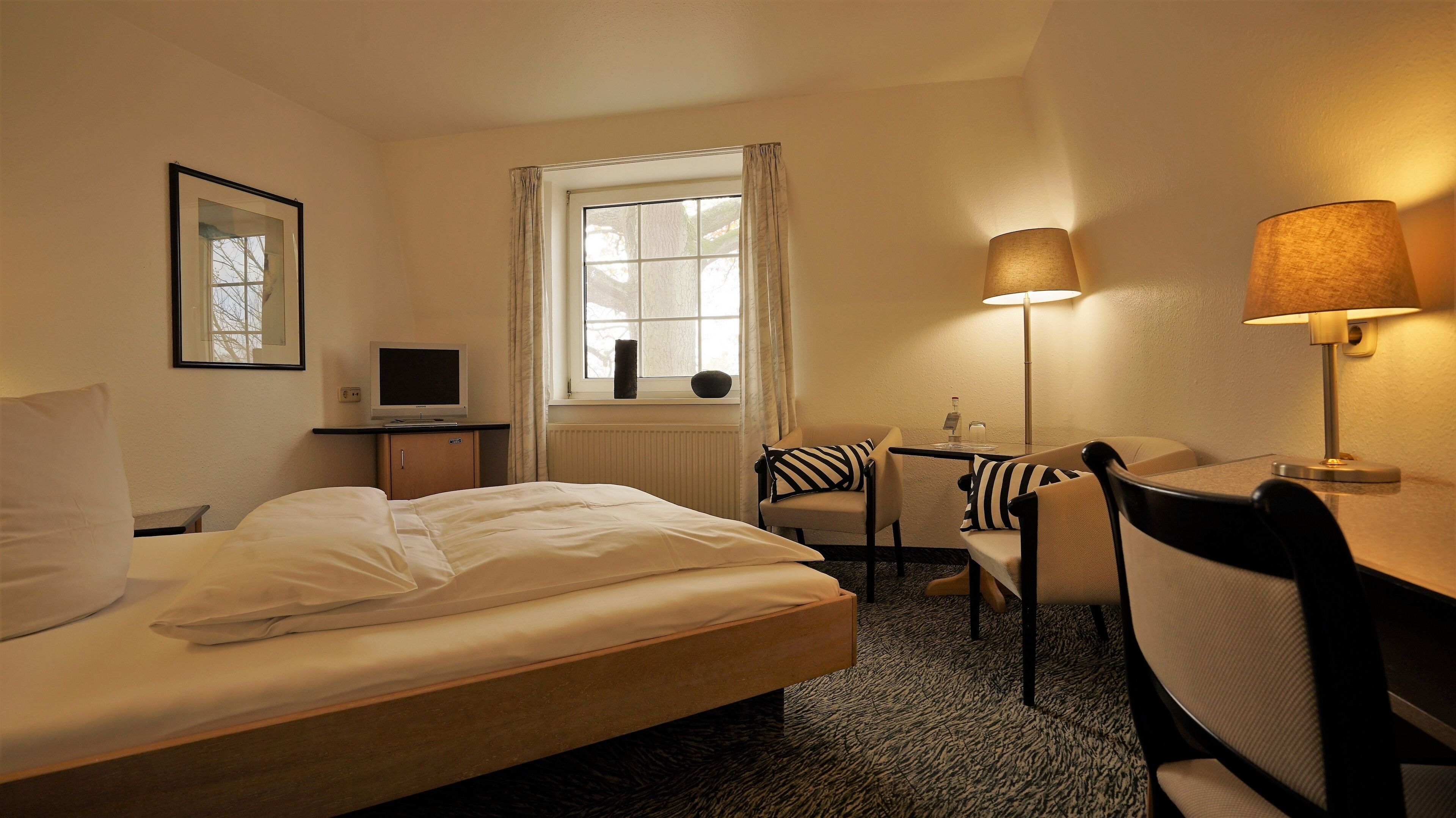 A hotel room with a bed, a desk, a chair, a lamp, a TV, and a window with curtains.