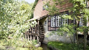 Ein traditionelles Haus mit einer Holztreppe, die zu einem Steineingang führt, umgeben von Grünpflanzen und einem kleinen Bach.