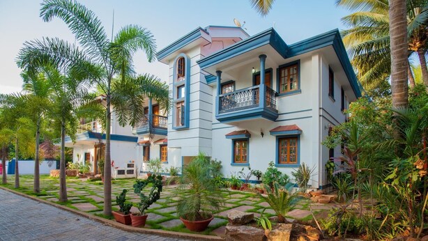 A two-story building with a balcony, surrounded by a garden with palm trees and potted plants. - Mistral Holidays (Benaulim)
