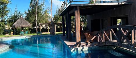 A swimming pool with a wooden deck and lounge chairs, surrounded by palm trees and a thatched-roof structure.