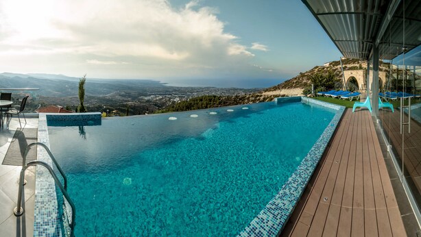 Une piscine à débordement à l’eau d’un bleu cristallin, une terrasse en bois et une vue sur une ville lointaine et des collines.